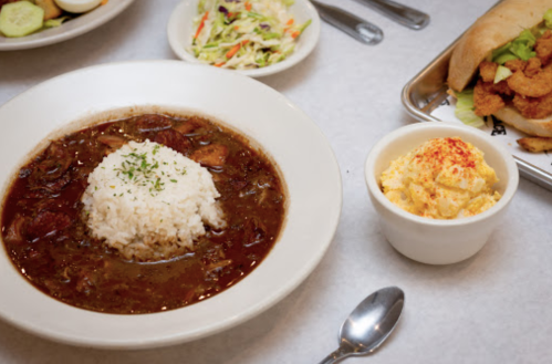 A bowl of stew with rice, a side of coleslaw, and a cup of creamy potato salad on a table.