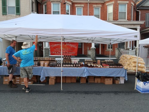 A vendor tent at a market displays jars of maple products, with two people chatting nearby.