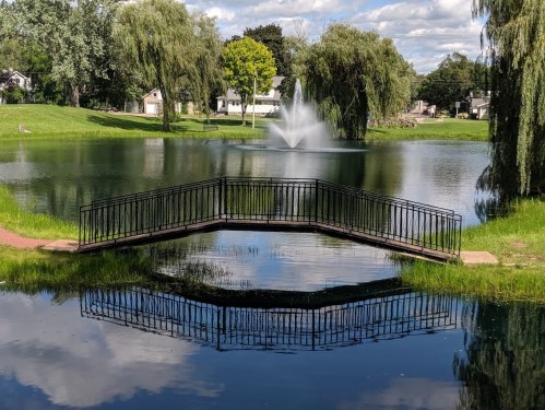 A serene pond with a fountain and a small bridge, surrounded by lush greenery and trees reflecting in the water.
