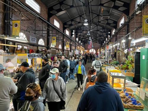 A bustling market scene with people browsing stalls, featuring food vendors and colorful displays in a large indoor space.