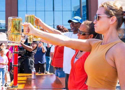 A group of people holding up large beer mugs at an outdoor event, with a crowd in the background.