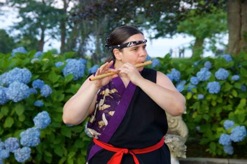 A person playing a flute in a garden surrounded by blue hydrangeas, wearing traditional attire with a headband.