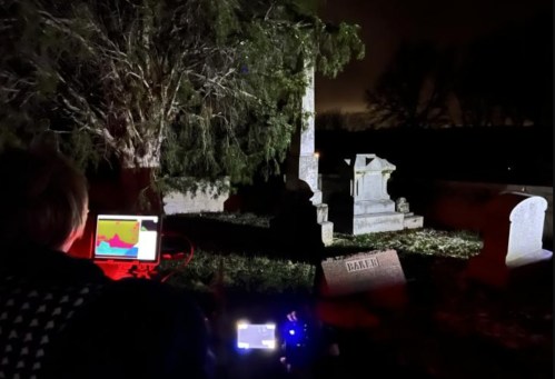 A person uses equipment in a dark cemetery, illuminated by red light, near a gravestone and a large tree.