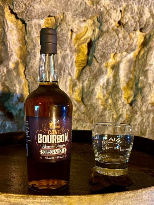 A bottle of Cave Bourbon whiskey next to a glass, set against a rustic stone background.