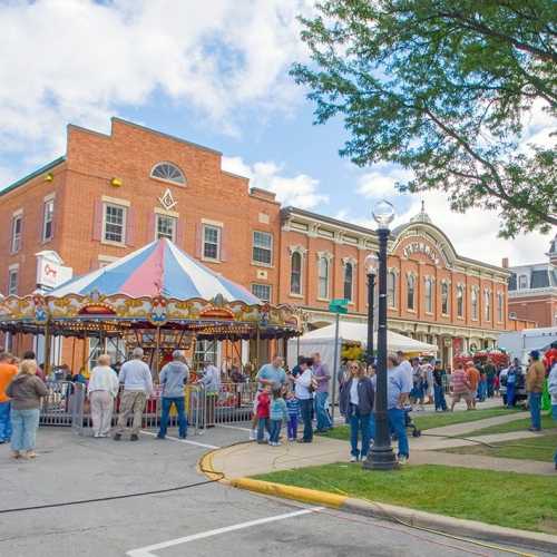 A lively outdoor fair scene with a carousel, people mingling, and historic buildings in the background under a blue sky.