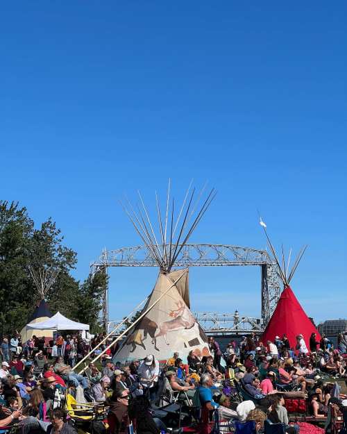Crowd gathered near colorful tipis under a clear blue sky, with a bridge visible in the background.