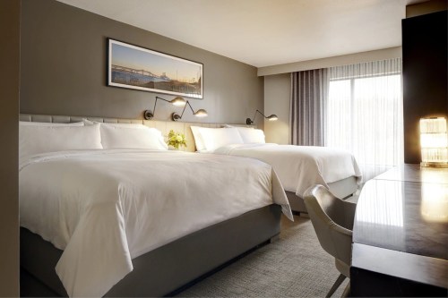 A modern hotel room featuring two queen beds, a desk, and large windows with natural light.