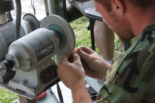 A person in a camouflage shirt sharpens a tool on a grinding machine outdoors.