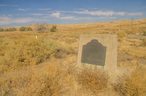 A weathered historical marker stands in a dry, grassy landscape under a clear blue sky.