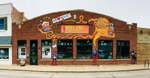 Colorful mural on a brick storefront featuring movie-themed art, with "Reel Art" sign and playful characters holding popcorn.