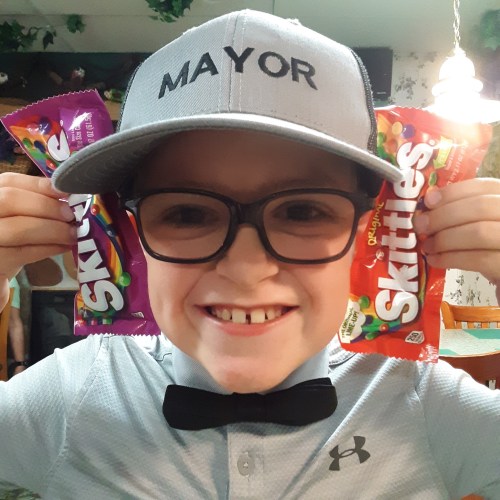A smiling child wearing a "MAYOR" cap and bow tie holds two packs of Skittles, one purple and one red.