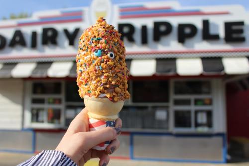 A hand holds a colorful ice cream cone topped with sprinkles and nuts in front of a Dairy Ripple shop.