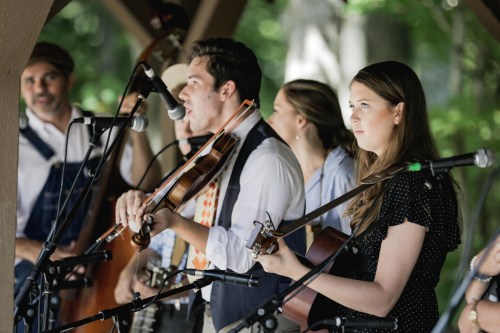 A group of musicians performs on stage, featuring a violinist and a guitarist, surrounded by greenery.