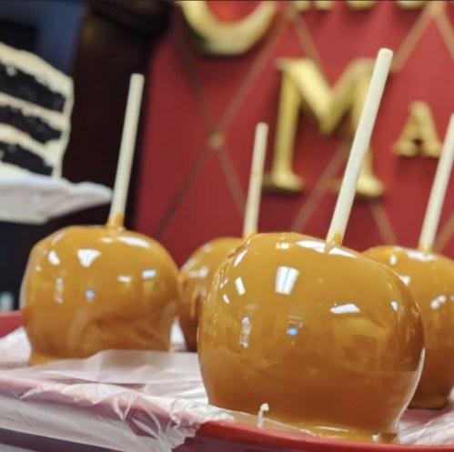 A tray of shiny caramel apples on sticks, with a blurred cake in the background and a decorative sign.