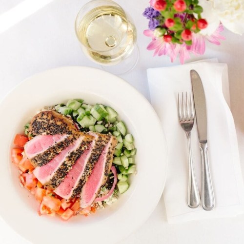 A plate of seared tuna on a bed of diced vegetables, accompanied by a glass of white wine and a neatly set table.