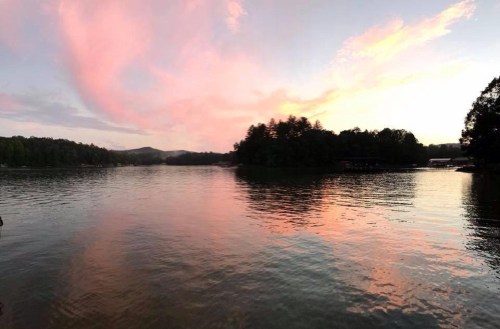 A serene lake at sunset, reflecting pink and orange clouds with trees lining the shore.