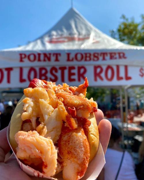 A hand holding a lobster roll filled with chunks of lobster, with a food stall in the background.