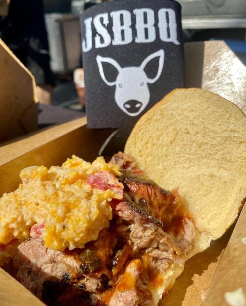 A pulled pork sandwich with mac and cheese, served in a takeout box, with a JSBBQ logo in the background.