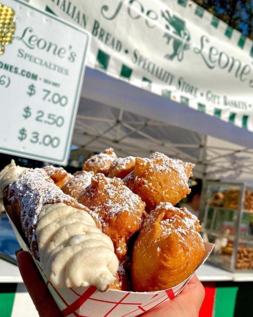 A hand holding a tray of fried pastries dusted with powdered sugar, with a sign for Joe Leone's in the background.