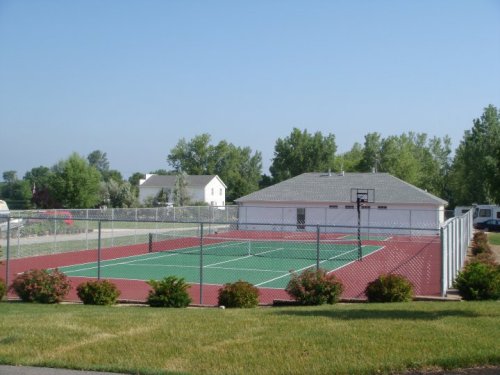 A fenced tennis court with a basketball hoop, surrounded by greenery and a residential building in the background.