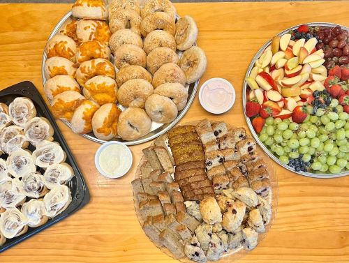 A spread of pastries, bagels, fruit, and desserts arranged on platters on a wooden table.