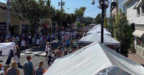A bustling street fair with crowds of people and vendor tents lining the road under clear blue skies.