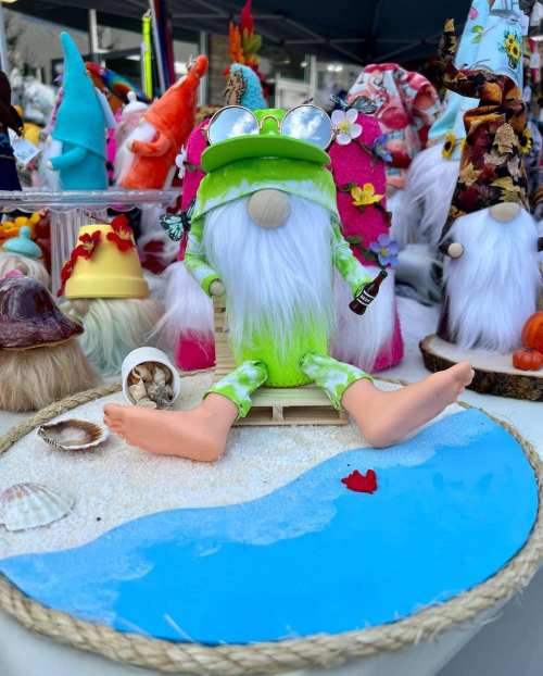 A whimsical gnome with a green outfit and sunglasses relaxes on a sandy beach scene, holding a drink.