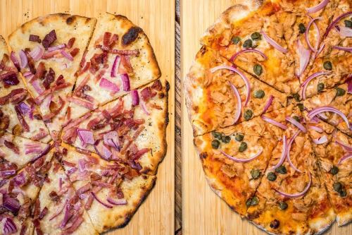 Two pizzas on a wooden board: one topped with bacon and red onions, the other with chicken, red onions, and capers.