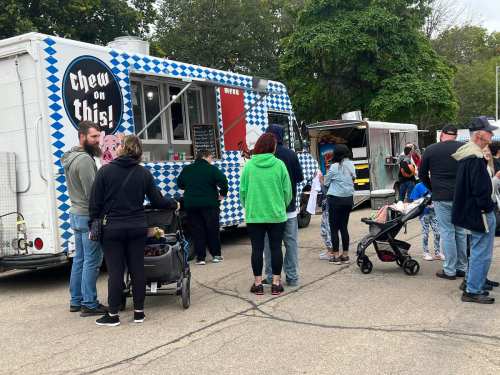 A food truck with a checkered design serves a crowd of people, some with strollers, at an outdoor event.