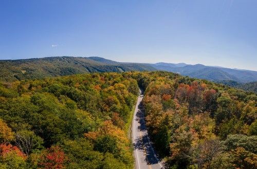Aerial view of a winding road through vibrant autumn foliage and rolling hills under a clear blue sky.