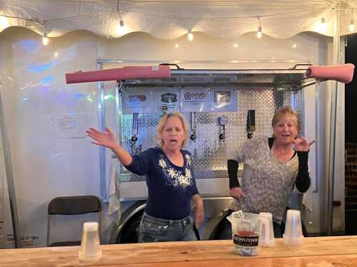 Two women joyfully dancing behind a bar with cups and a beer tap, under string lights in a festive setting.
