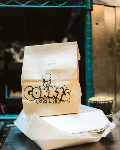 A takeout bag from Corky's Ribs & BBQ sits on a table next to a white food container.