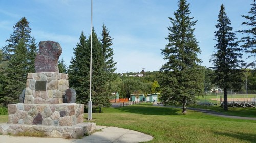 A stone monument stands in a park surrounded by trees, with a baseball field and buildings visible in the background.