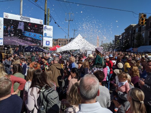 A large crowd gathers at an outdoor event, with a screen displaying images and bubbles floating in the air.