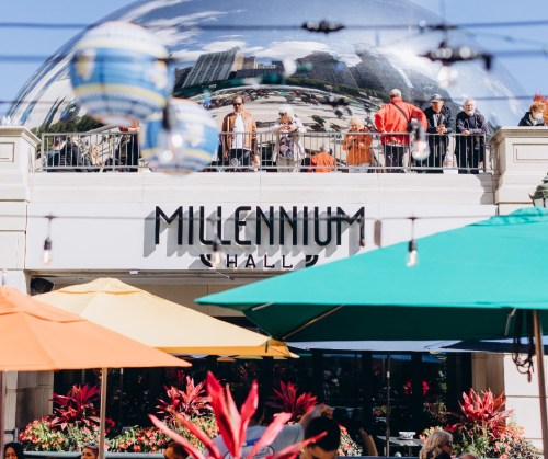 A vibrant outdoor scene at Millennium Hall, featuring colorful umbrellas and people on a balcony reflected in a dome.