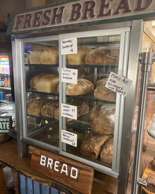 A display case filled with various types of fresh bread, labeled with prices and a sign asking for assistance.
