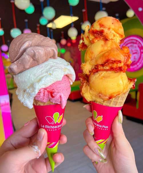 Two colorful ice cream cones held in hands, featuring chocolate, strawberry, and mango flavors, set in a vibrant shop.
