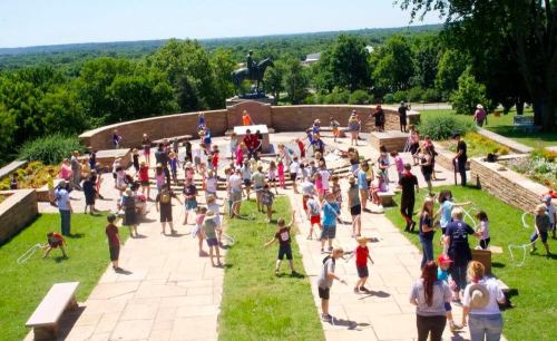 A large group of children and adults play with hula hoops on a sunny day in a park, with greenery and a statue in the background.