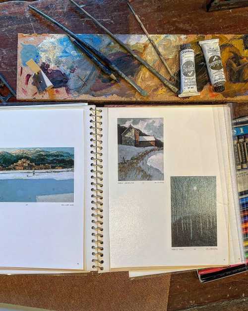 An open sketchbook on a wooden table, featuring art prints, paint tubes, and brushes scattered around.