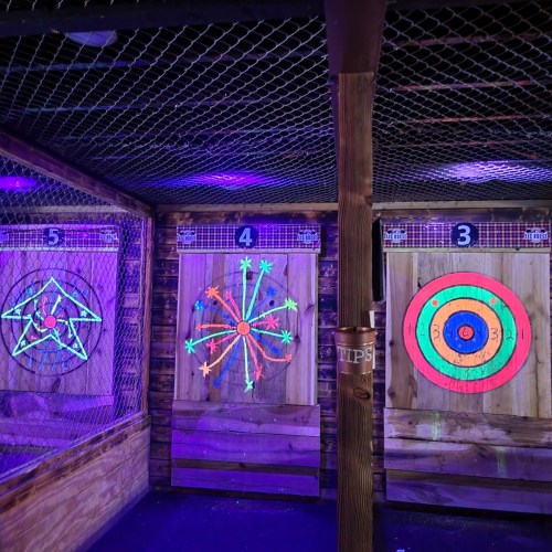 A dimly lit indoor space with three colorful dartboards, each featuring unique designs and illuminated by purple lights.