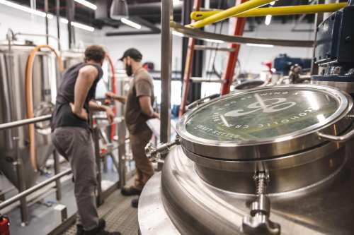 Two brewers discuss equipment in a brewery, with a focus on a shiny brew kettle in the foreground.