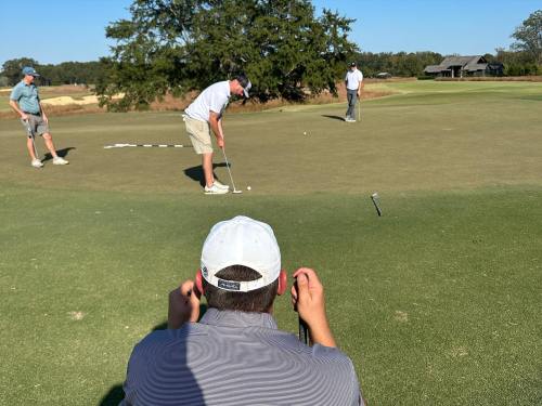A golfer putts on a green while two others watch from a distance, with trees and a building in the background.
