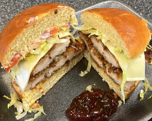 A close-up of a sandwich with layers of fried chicken, lettuce, cheese, and tomato, served with a side of sauce.