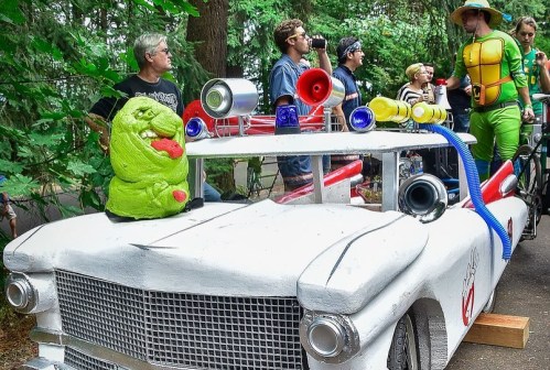A creatively decorated car with colorful speakers, featuring a green monster figure on the hood, surrounded by people in costumes.
