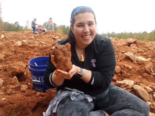 A smiling woman holds a large rock while sitting on a dirt mound, with people and trees in the background.