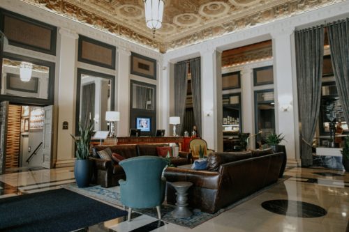 Elegant hotel lobby featuring plush seating, ornate ceilings, and stylish decor with large windows and plants.