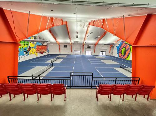 A spacious indoor sports facility with blue tennis courts and vibrant orange walls, featuring red spectator seating.
