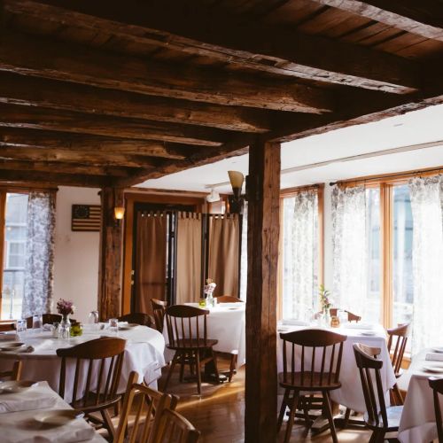 Cozy restaurant interior with wooden beams, white tablecloths, and natural light from large windows.