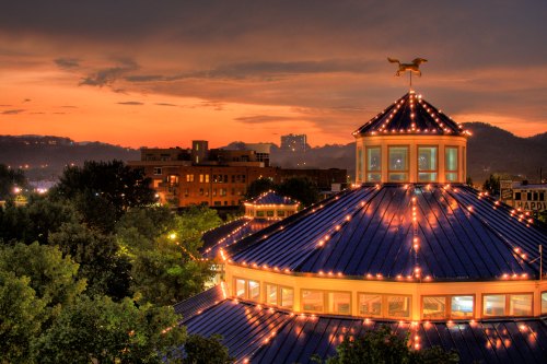 A rooftop view of a circular building adorned with lights at sunset, surrounded by trees and distant city skyline.