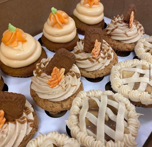 A box of assorted cupcakes with cream frosting, decorated with orange flowers and cookie pieces.